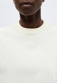 Κοντινό ενός ατόμου που φοράει ένα εκρού μπλουζάκι με στρογγυλή λαιμόκοψη και ανάγλυκη λέξη "BORG" στο κέντρο του στήθους.