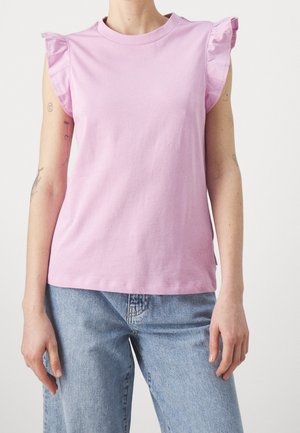 Osoba ubrana w jasnofioletowy bezrękawnik z falbankami na ramionach oraz jasnoniebieskie dżinsy, stojąca na prostym tle.