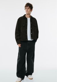 Jonge man in een zwarte jas, witte t-shirt, losse zwarte broek en witte sneakers, staand met één hand in zijn zak en opzij kijkend.