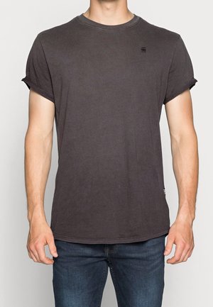 Homme portant un t-shirt gris foncé uni à manches courtes avec les manches retroussées et un jean bleu, debout devant un fond uni.
