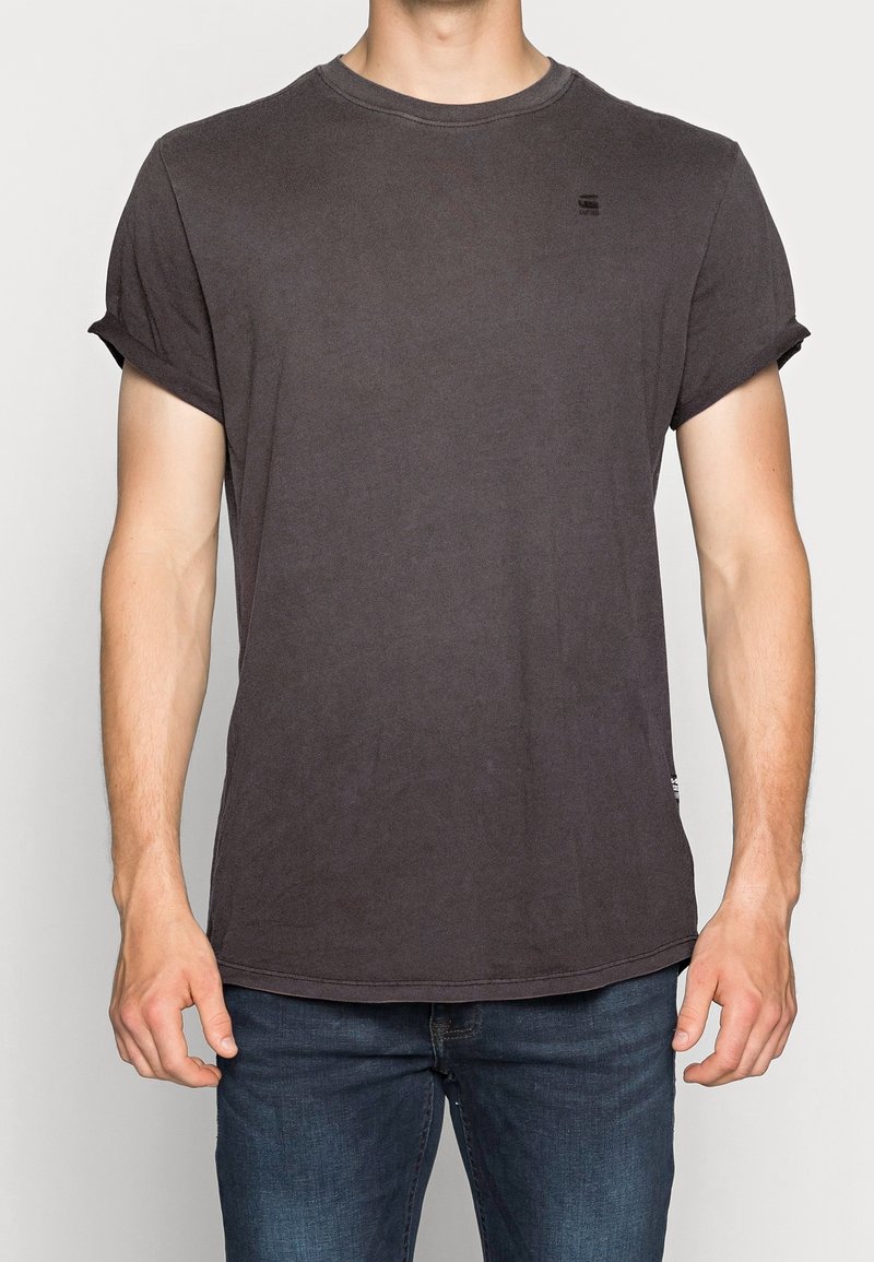 Homme portant un t-shirt gris foncé uni à manches courtes avec les manches retroussées et un jean bleu, debout devant un fond uni.
