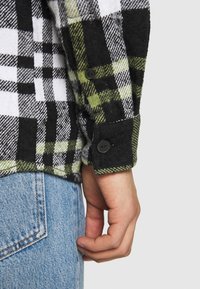 Camicia in flanella con un motivo a quadri nero, bianco e verde. Presenta un polsino con bottone nero ed è realizzata in un tessuto morbido e testurizzato.