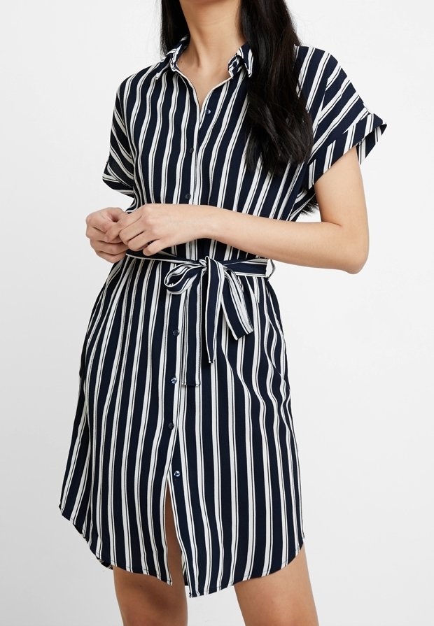 Femme portant une robe chemise à manches courtes à rayures verticales bleu marine et blanc, avec une ceinture nouée à la taille et une fermeture à boutons devant.