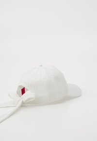 Gorra de béisbol blanca con tejido texturizado, visera curvada y un gran lazo en la parte trasera. Presenta un logo bordado en la parte frontal.