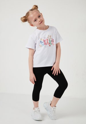 Noorblondide patsidega noor tüdruk, kes kannab valget t-särki, millel on kiri "HELLO", musti kapri-pükse ja valgeid tossusid, seisab ühe jala kõverdatuna.