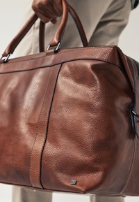 Brun lædertaske med en kornet struktur, udstyret med robuste håndtag, kontrasterende syninger og en lille logodetalje på siden.