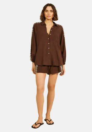 Kobieta w luźnej brązowej koszuli zapinanej na guziki i dopasowanych szortach z białym kwiatowym haftem na rękawach i bokach, czarne klapki, stoi.