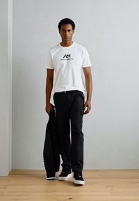 T-shirt en coton blanc avec logo noir, associé à un pantalon cargo noir et des baskets noires. Le modèle tient une veste noire, debout sur un sol en bois.