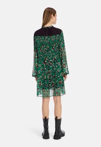Grünes Leopardenmuster-Kleid mit transparenten langen Ärmeln, schwarzem Einsatz und smockierter Taille. Getragen mit schwarzen Knöchelstiefeln.