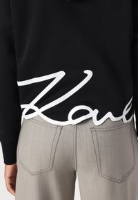 Schwarzer Sweatshirt mit weißem, gesticktem "Love"-Text am Saum, kombiniert mit hellgrauen, strukturierten Hosen mit Gesäßtaschen.