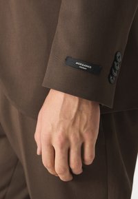Suknja smeđe odijela sa vidljivom etiketom na kojoj piše "JACK & JONES", četiri crna dugmeta i glatka tekstura tkanine.
