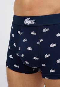 Σκούρα μπλε εσώρουχα τύπου μποξέρ με λευκά λογότυπα κροκόδειλου, φορεμένα από ένα άτομο, που δείχνει την περιοχή της κοιλιάς και το επάνω μέρος των μηρών.