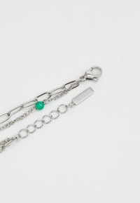 Sidabrinis metalinis apyrankė, kurią sudaro įvairios grandinėlės, žalia spalvos perlas ir stačiakampis ženklelis su įspaudu "LACOSTE". Įtrauktas užsegimas.