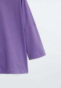 Top viola a maniche corte in tessuto liscio con una vestibilità leggermente ampia e cuciture visibili lungo le maniche e l'orlo.