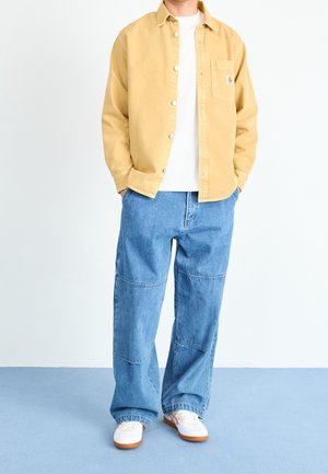 Personne portant une veste jaune à boutons, une chemise blanche, un jean bleu ample et des baskets blanches debout sur un sol bleu clair.