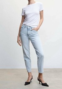 Femme portant un t-shirt blanc à manches courtes, un jean taille haute bleu clair et des talons slingback noirs, debout devant un fond uni.