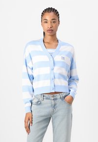 Modra in bela prugasta jopica z gumbnim zapiranjem spredaj, v-izrezom in rebrastimi manšetami, v kombinaciji s svetlim denim džinsom.