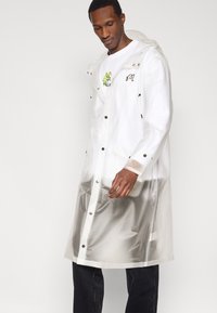 Διαφανές αδιάβροχο από PVC με λευκές λεπτομέρειες, κουμπώματα με τσιμπιδάκια και δύο μπροστινές τσέπες. Διαθέτει κουκούλα και κεντητό σχέδιο στο στήθος.