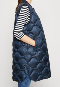 Námořnický modrý prošívaný puffer vest s vlnitým vzorem, bočními kapsami a vysokým límcem. Nositý přes pruhovanou košili s dlouhým rukávem.