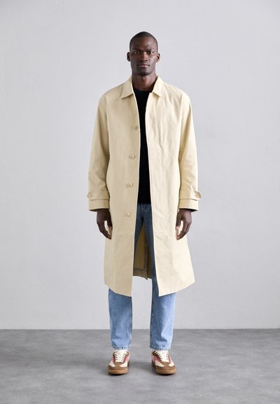 Manteau long beige avec col et fermeture à boutons, porté sur une chemise noire et un jean bleu. Les chaussures présentent un design multicolore et une semelle marron.