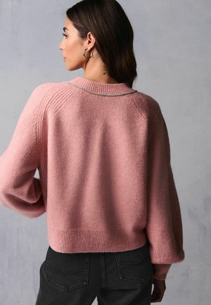 Rosa gestrickter Pullover mit strukturierter Oberfläche, gerippten Bündchen und einem runden Halsausschnitt, verziert mit metallischen Kettendetails. Schwarze Jeans runden den Look ab.