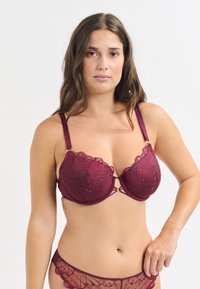 Reggiseno in pizzo borgogna con motivo floreale, supporto con ferretto, bordi scallop e spalline regolabili. Caratterizzato da un dettaglio centrale decorativo.