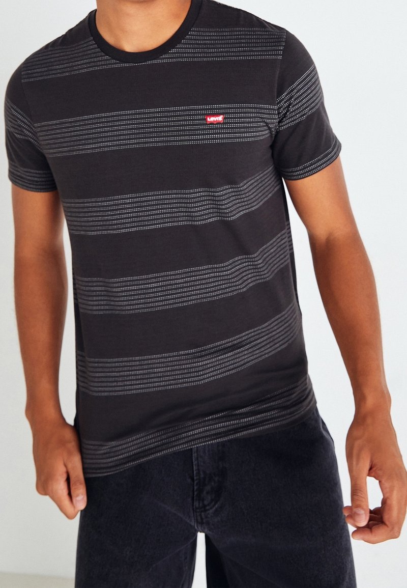 Homme portant un T-shirt Levi's noir à manches courtes avec des rayures horizontales grises à pois et un pantalon foncé, debout devant un fond clair.