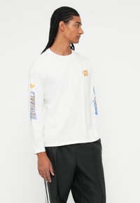 Vyras su pintomis plaukais, vilkintis baltą ilgą rankovę Adidas Originals marškinėlį su oranžiniais ir mėlynais logotipais bei juodas kelnes su baltomis juostelėmis.