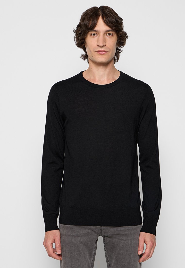 Schwarzer Strickpullover mit langen Ärmeln und Rundhalsausschnitt, der eine glatte Textur und eine figurbetonte Passform aufweist. Kombiniert mit grauen Hosen.
