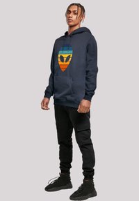 Navy hoodie with a front graphic featuring a stylized alien face in orange, yellow, and blue stripes. Pockets and ribbed cuffs included.