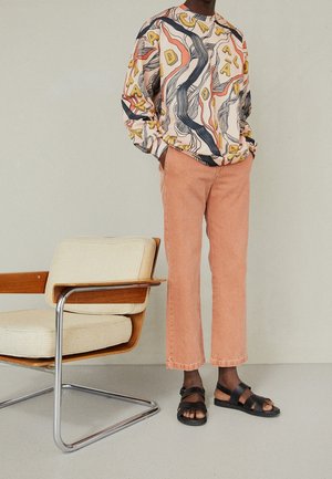 Person trägt ein gemustertes langärmeliges Hemd, pfirsichfarbene Hose und schwarze Sandalen und steht neben einem beigen, gepolsterten Holzstuhl.