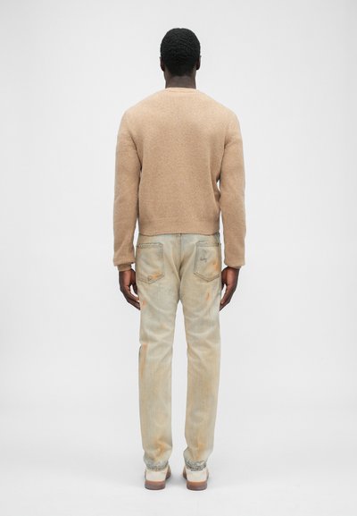 Beige gestrickter Pullover mit einem gerippten Rundhalsausschnitt, kombiniert mit hellblauen, verwaschenen Jeans, die mit Farbklecksen verziert sind.