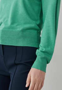 Pull vert en maille avec poignets et ourlet côtelés, associé à un pantalon bleu marine. Le pull présente une texture lisse et une coupe ajustée.