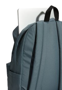 Szary plecak z wyściełanymi paskami i boczną kieszenią, w której znajduje się srebrny laptop. Tkanina o teksturowanej powierzchni z gładkim wykończeniem oraz czarną pętlą do zawieszania.