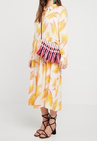 Abito a maniche lunghe color crema con motivo floreale e palme gialle e arancioni. Presenta una borsa a tracolla intrecciata con pompon rosa e rossi. Sandali neri con cinturini.