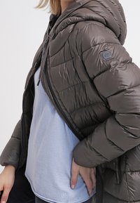 Donkergrijze pufferjack met een capuchon, voorzien van een gestikt patroon. Draagt open over een lichtblauwe fleecetop, wat een casual gelaagde stijl laat zien.