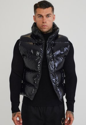Uomo con capelli corti e scuri che indossa un gilet imbottito nero lucido sopra un maglione nero, in piedi davanti a uno sfondo grigio uniforme.