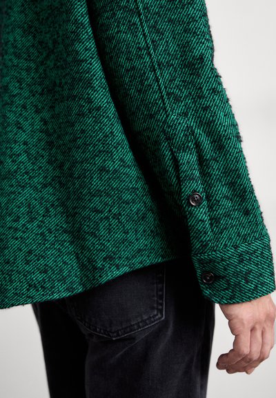 Chemise verte à motif chevrons, avec un tissu texturé, des poignets boutonnés et un ourlet légèrement arrondi, associée à un jean foncé.