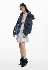 Üle suurusega tumehall bomberjakk karvaga vooderdatud kapuutsiga, kantud helehalli kleidi peale. Koos läikivate mustade pahkluu saapadega ja hoides mustrilist kotti.