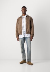 Brown bomber jacket with elastic cuffs, white striped shirt, light faded jeans, and brown slip-on shoes. Clean and minimal design.