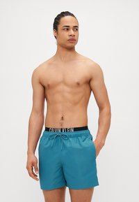 Τελικά σορτς μαγιό σε απόχρωση τιρκουάζ από ελαφρύ υλικό, με ελαστική ζώνη στη μέση και μαύρο λογότυπο Calvin Klein, καθώς και ρυθμιζόμενο κορδόνι.