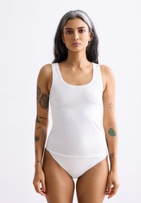 Weißes Tanktop und Bikini-Set, gefertigt aus einem weichen, glatten Stoff. Verfügt über einen runden Ausschnitt und breite Träger. Tätowierungen sind auf den Armen sichtbar.