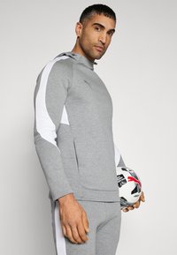 Grå atletisk hoodie med hvide accenter, udstyret med hætte med lynlås og sidelommer, båret med matchende grå bukser; holder en mønstret fodbold.