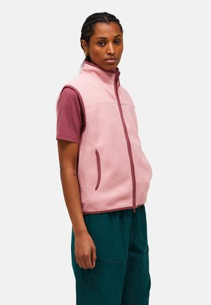 Woman with braided hair wearing a pink fleece zip-up vest over a mauve short-sleeve shirt and teal pants, facing forward with a neutral expression.