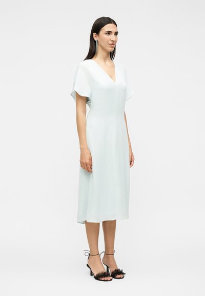 Γυναίκα στέκεται φορώντας λευκό μίντι φόρεμα με V-λαιμόκοψη και φαρδιά μανίκια, και μαύρα σανδάλια με τακούνι, κορδέλες στον αστράγαλο και φτερά ως διακοσμητικά.