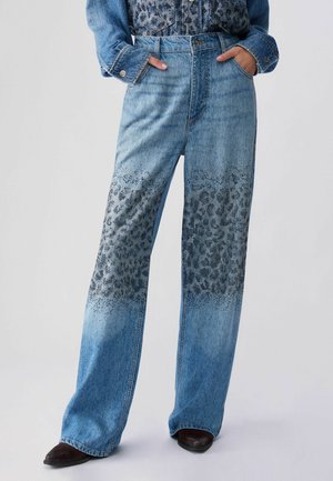 Persoon staat met de handen in de zakken, draagt een blauwe high-waisted wide-leg spijkerbroek met zwart luipaardpatroon op de dij- en kniegebieden.