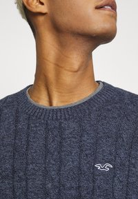 Pull en maille torsadée bleu marine avec un col rond côtelé et une bordure de col gris clair. Présente un petit logo blanc sur la poitrine.