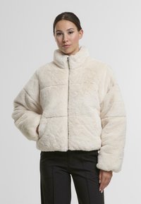 Veste en fausse fourrure crème avec un col haut, fermeture éclair et manches surdimensionnées, dotée d'une finition texturée et sans embellissements visibles.