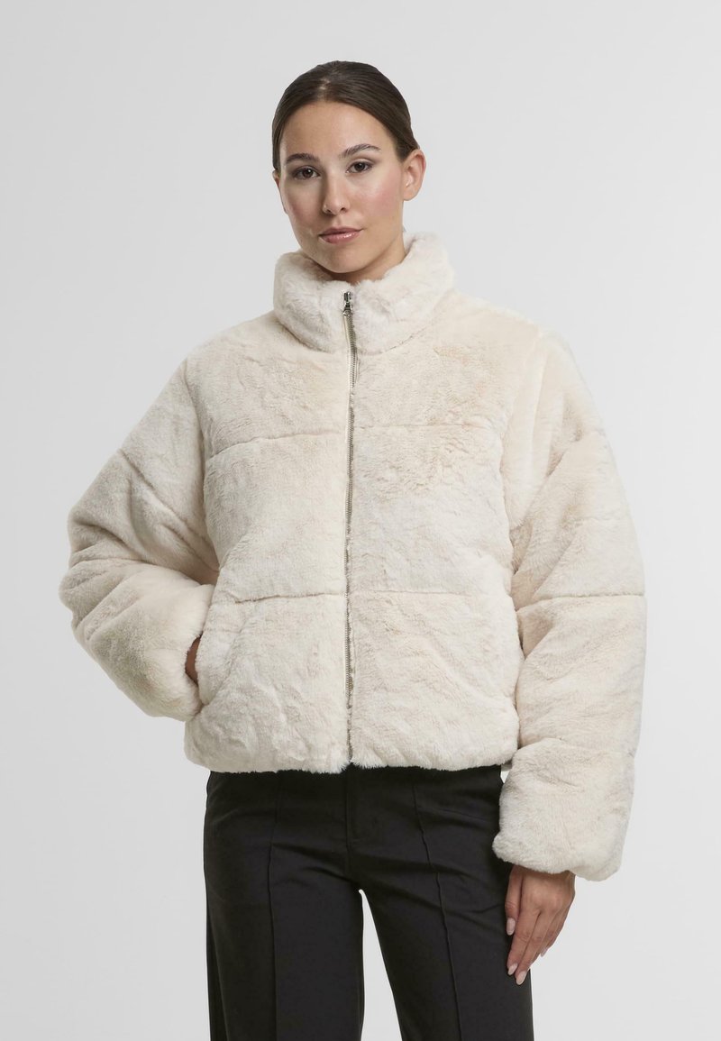 Veste en fausse fourrure crème avec un col haut, fermeture éclair et manches surdimensionnées, dotée d'une finition texturée et sans embellissements visibles.