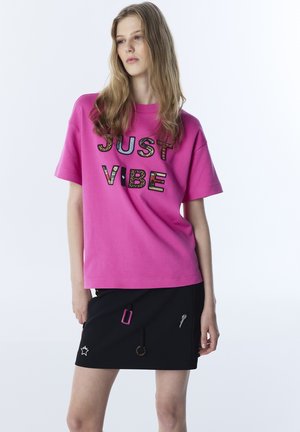Jonge vrouw die een felroze T-shirt met de tekst "JUST VIBE" draagt en een zwarte rok met decoratieve patches, poserend tegen een effen witte achtergrond.
